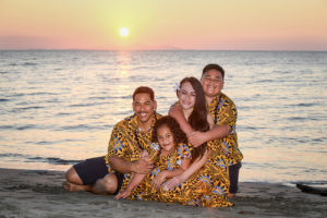 The family cuddles on the shore, behind, a golden Fiji sunset