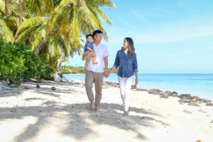 The family strolls on the white sand beach against green palm trees and blue sea