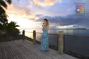 On a terrace a beautiful woman smelling the flavor of a glass of red wine in front of a impressive sunset in Fiji logo