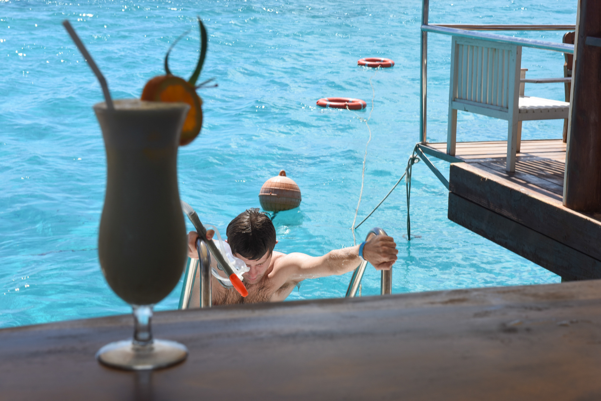 Composition with a glass of a cocktail in front of the turquoise sea and guests swimming and getting out the water