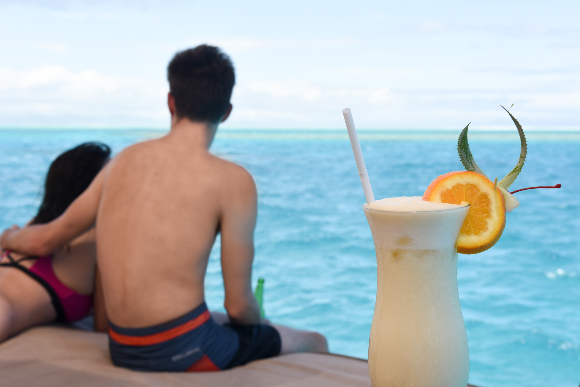 Composition with a glass of a cocktail in front of the turquoise sea and couple relaxing on the boat