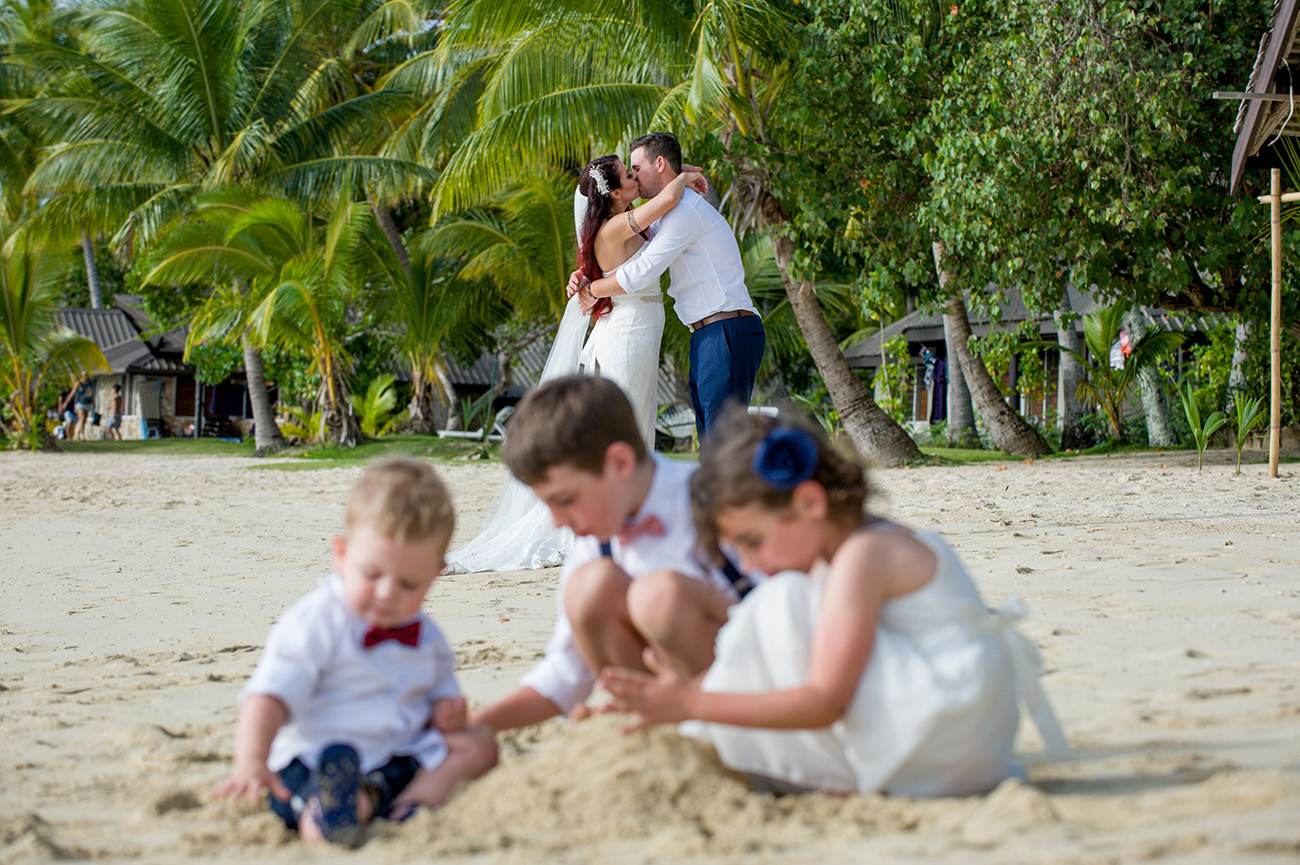 Newly married couple kiss in the background as children play with sand castles on the shore