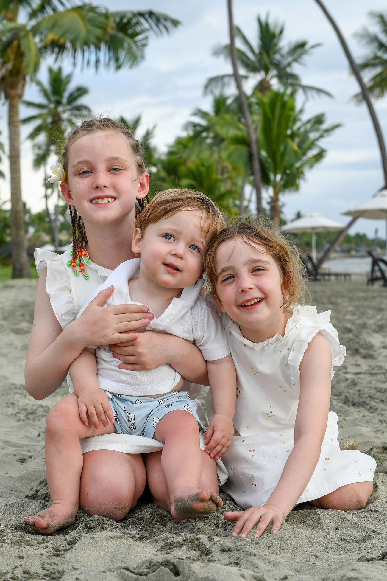 Sister, sister and brother in white cuddle against palm trees